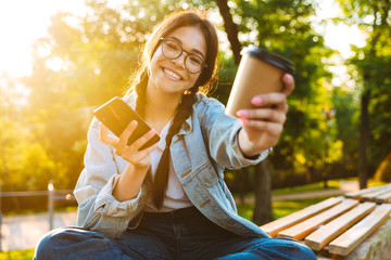 Wall Mural - Happy cute young student girl wearing eyeglasses sitting outdoors in nature park using mobile phone drinking coffee.