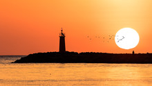 Romantic Sunrise Over The Mediterranean In The Spanish Port City Denia. A Man Looks Into The Sun, The Lighthouse Flashes And A Flock Of Birds Fly By.
