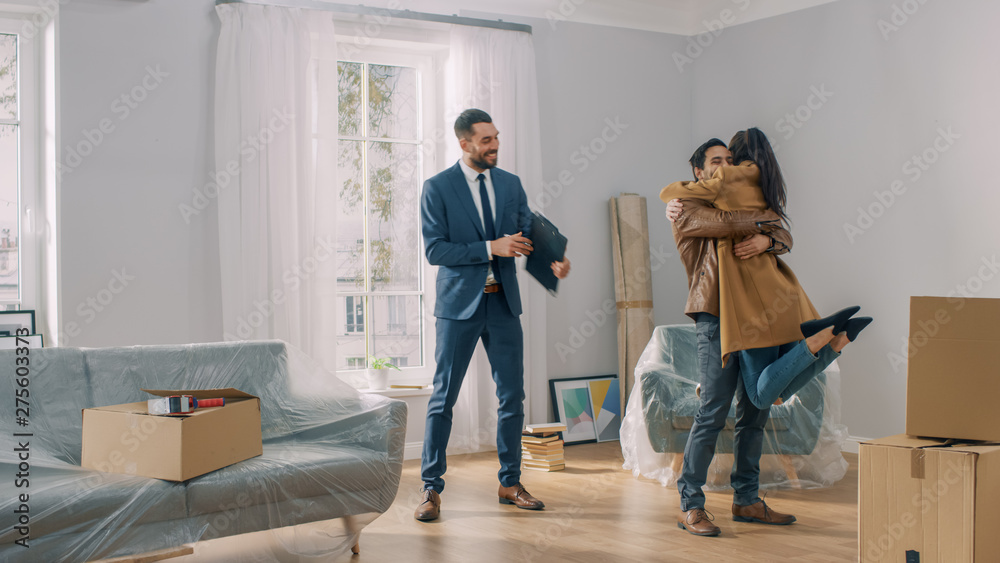 Real Estate Agent Shows Bright New Apartment to a Young Couple. Successful Young Couple Becoming Homeowners. Girl Jumps Into His Boyfriend's Arms Hug. Spacious Bright Home with Big Windows. - obrazy, fototapety, plakaty 