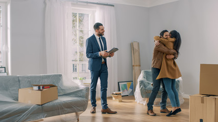 Real Estate Agent Shows Bright New Apartment to a Young Couple. Successful Young Couple Becoming Homeowners, Embraces and Hugs Each Other. Spacious Bright Home with Big Windows.