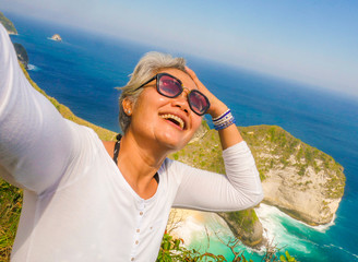 middle age 50s happy and cheerful Asian woman with grey hair taking selfie with mobile phone at beautiful tropical beach island smiling at cliff viewpoint enjoying Summer