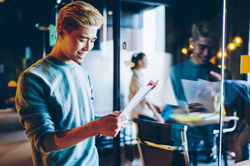 Positive asian male student reading report satisfied with good job, prosperous handsome male employee in casual wear checking information from paper enjoy startup incomes for company in office.