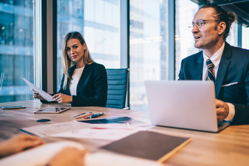 Successful adult female entrepreneur sitting at table in office company with paper document in hand and looking at camera during brainstorming meeting with corporate colleagues. Entrepreneurs people