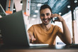 Portrait of positive male freelancer sitting at desktop with modern laptop computer and smiling at camera while enjoying smartphone call communication with friend using 4g internet connection
