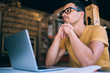 Contemplative young male student in spectacles for vision correction looking away and pondering thinking on idea for university project, pensive hipster guy sitting at desktop with modern laptop