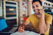 © BullRun - Portrait of happy hipster guy looking at camera and smiling while sitting at desktop with laptop computer for e learning and textbook for knowledge education, cheerful male student feeling good