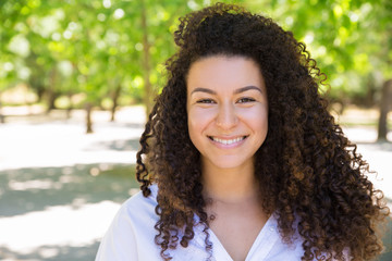 Happy young curly-haired lady posing at camera in park. Beautiful lady wearing blouse and looking at camera with green trees in background. Woman portrait concept. Front view.