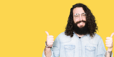 Young hipster man with long hair and beard wearing glasses success sign doing positive gesture with hand, thumbs up smiling and happy. Looking at the camera with cheerful expression, winner gesture.