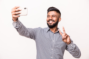 Canvas Print - Young indian young man with beard and mustache taking selfie using smartphone make peace gesture, isolated over white background