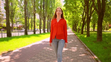 Poster - Nice Red Haired Woman In Trendy Clothes Goes By Confident Stride On Park Path. Green Trees On Background.
