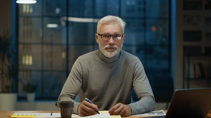 Poster - Portrait of the Handsome and Successful Middle Aged Bearded Businessman Working at His Desk Using Laptop Computer. Smiles at Camera. Working at Night from Cozy Home Office / Studio with Window
