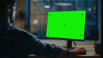 Poster - Over the Shoulder: Creative Young Man Sitting at His Desk Using Desktop Computer with Mock-up Green Screen. Evening in the Stylish Office Studio with City Window View