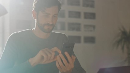 Wall Mural - Professional Creative Man Sitting at His Desk in Home Office Studio Using Smartphone Man Using Mobile Phone, Browsing Through Internet, Social Networks / Media, Working on Laptop. Camera Moving Around