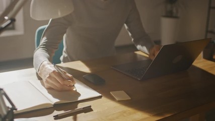 Poster - Anonymous Creative Man Sitting at His Desk in Home Office Studio Working on a Laptop Computer, Writes down Notes. Low Angle 360 Degree Tracking Arc Shot Movement. Camera Moving Around