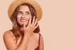 © sementsova321 - Photo of attractive smiling woman with long hair, has happy facial expression, applaying sunscreen, wearing straw hat, wanting to tan, isolated on beige wall. Summertime, vacation, sunscreen concept.