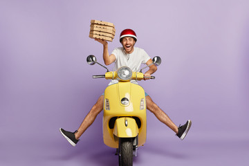 Busy deliveryman being in hurry, carries cardboard boxes with pizza, delivers to customers, poses on yellow scooter, wears helmet, white t shirt and sportshoes, spreads legs, has happy expression