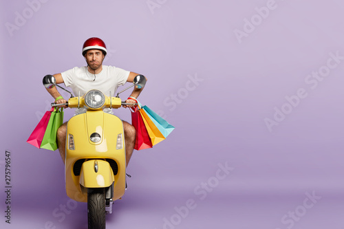Confident busy man delivers online order for customer, rides fast motorbike, carries five colorful bags with products, has excellent driving skills. Shopaholic returns home with shopping bags