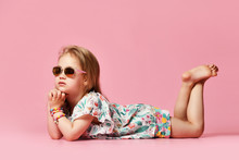 Little Girl Child Looks To The Side Lying On The Floor Of The Studio In Summer Clothes In Sunglasses On A Pink Background One