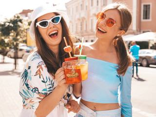 Two young beautiful smiling hipster girls in trendy summer clothes and panama hat.Sexy carefree women posing in the street.Positive models having fun in sunglasses.Drinking fresh cocktail smoozy drink