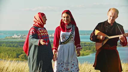 Wall Mural - People in traditional russian clothes walking on the field and singing a song - a man playing balalaika