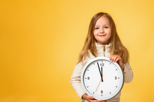 Cute Little Girl In A Jacket Holds A Big Clock On A Yellow Background. The Concept Of Education, School, Deadlines, Time To Study, Autumn. Copy Space