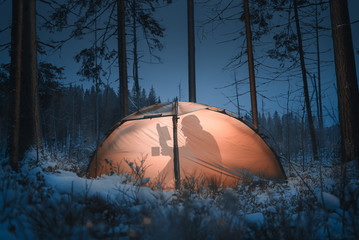 Silhouette of a man in a tent. He reads a book and drinks tea. Winter time. Pine forest