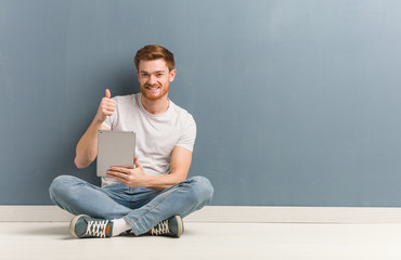 Young redhead student man sitting on the floor smiling and raising thumb up. He is holding a tablet.