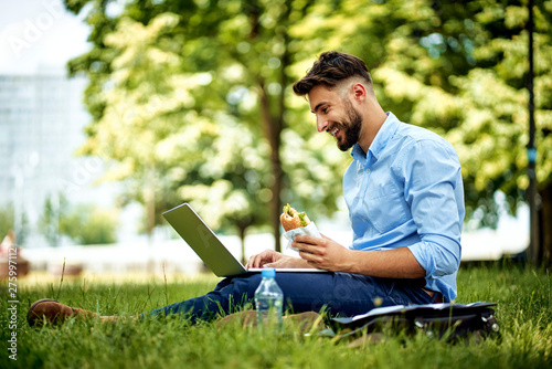 Fotografía Young cheerful businessman sitting in the park with sandwich and laptop
