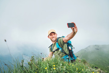 Wall Mural - smiling backpacker man taking selfie picture using smartphone and showing thumbs up during walking b