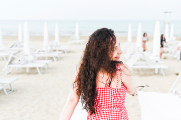 A woman in a red dress and curly hair is standing on the beach. A man with happy emotions. Walk along the sea promenade. Travel and tourism in the summer. Copy space