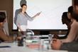 Positive attractive young female analyst in stripped blouse standing against projection screen and pointing at graph while explaining sales data to colleagues at meeting