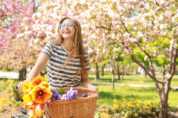 Beautiful young woman with bicycle and flowers in park on spring day