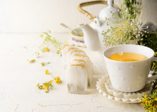 White Tea Cup With Tea Bags Of Herbal Tea Standing  On White Table With Teapot And Fresh Medical Herbs And Flowers. Close Up. Healthy Preventive Drink Treatment