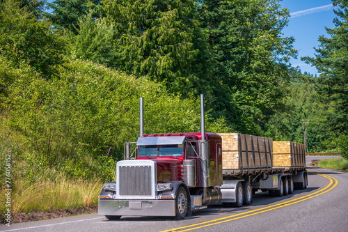 Big rig red semi truck with classic style transporting lumber on flat bed semi trailer driving on winding forest road