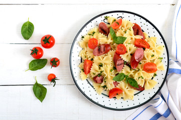 Wall Mural - Tasty pasta farfalle with grilled sausages, fresh cherry tomatoes and basil on a plate on a white wooden background. Top view, flat lay.