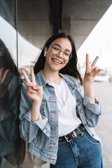 Sticker - Happy young pretty woman student wearing eyeglasses walking outdoors resting listening music with earphones showing peace gesture.