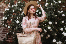 Outdoor Portrait Of Young Beautiful Fashionable Woman Wearing Wide Brim Straw Hat, Pink Dress, Holding Wicker Straw Basket Bag. Model Posing In The Blooming Rose Garden. Copy, Empty Space