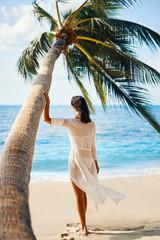 Wall Mural - Back view of pretty young woman relax and enjoy sea standing under palm tree on tropical beach