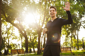 Handsome cheerful young sports fitness man standing in green park nature holding bottle with water waving to friends.