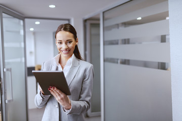 Wall Mural - Charming businesswoman in formal wear and with big toothy smile using tablet while standing in hall at work.