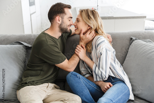 Photographie Smiling young loving couple indoors at home sitting on sofa hugging kissing