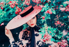 Young Woman With Creative Eye Makeup With Retro Arrow And Red Lips Looks Straight To Camera, Bright Colors Of Blooming Roses. Lady In Pink Elegant Dress With Wide-brimmed Hat And Black Ribbon Bow