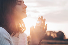 Teenager Girl Closed Her Eyes, Praying In A Field During Beautiful Sunset. Hands Folded In Prayer Concept For Faith, Spirituality And Religion. Peace, Hope, Dreams Concept