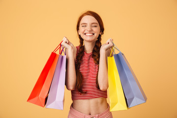 Sticker - Happy young beautiful redhead woman posing isolated over yellow background holding shopping bags.