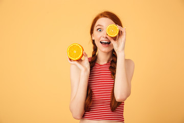 Canvas Print - Happy  young beautiful redhead woman posing isolated over yellow background holding orange.