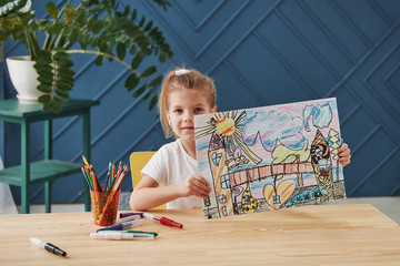 Nice interior in the room. Cute little girl in art school shows her first paintings drawn by pencils and markers