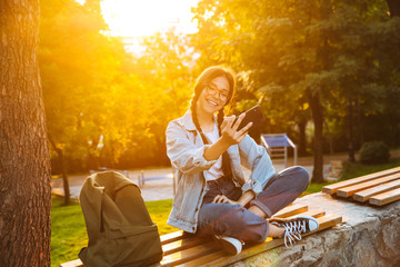 Canvas Print - Smiling laughing cute young student girl wearing eyeglasses sitting on bench outdoors in nature park with beautiful sunlight using mobile phone talking.