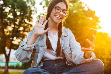 Poster - Happy cute young student girl wearing eyeglasses sitting on bench outdoors in nature park with beautiful sunlight talking by mobile phone waving.