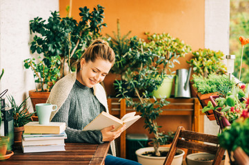 Wall Mural - Young beautiful woman relaxing on cozy balcony, reading a book, wearing warm knitted pullover, cup of tea or coffee on stack of books