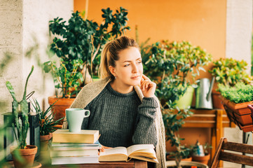 Young beautiful woman relaxing on cozy balcony, reading a book, wearing warm knitted pullover, cup of tea or coffee on stack of books